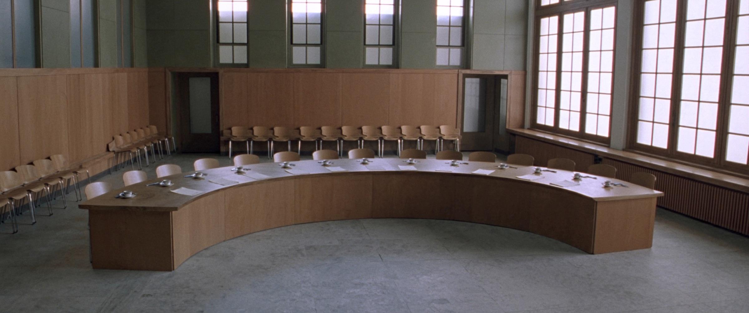 La salle du conseil d'une maison communale : une longue table, des rangées de chaises, hautes fenêtres et sol de pierre dans la lumière du jour.