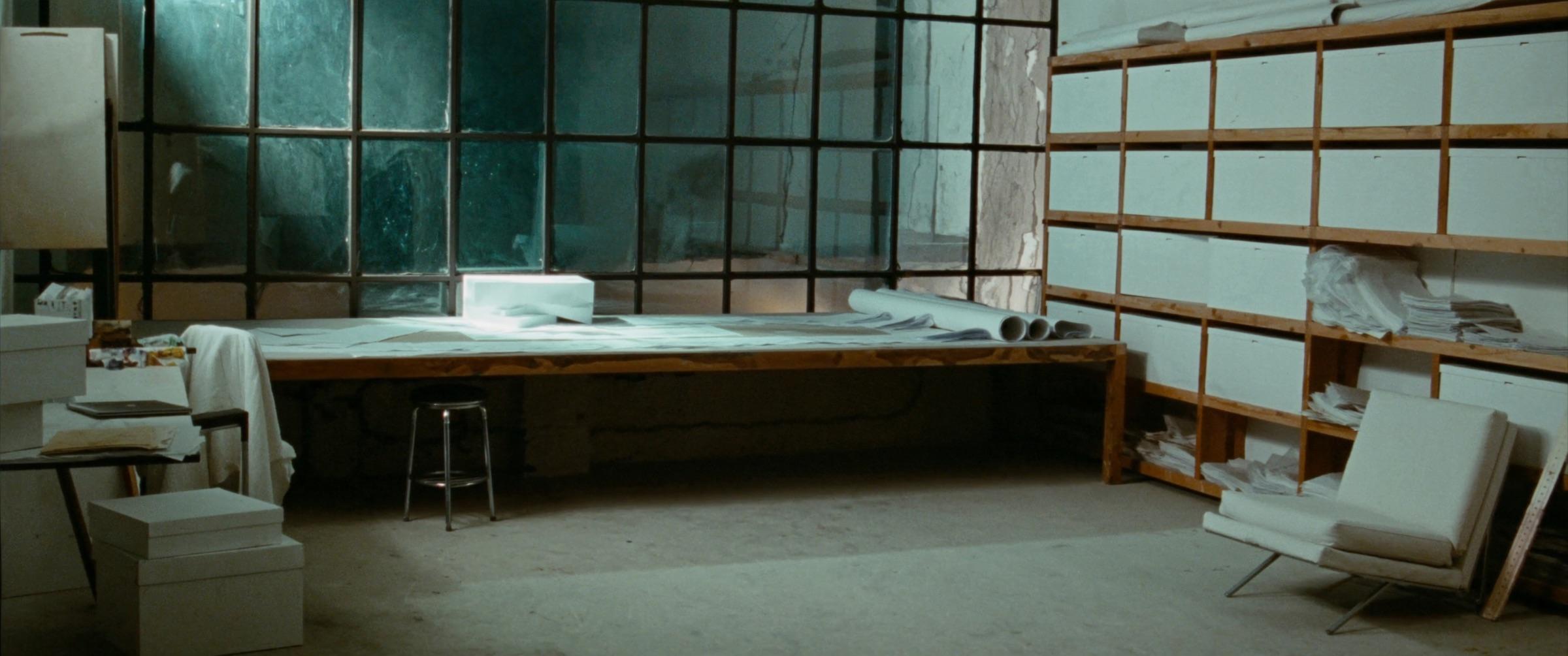 Un atelier d'architecture : une longue table à dessin, des plans roulés et une maquette, hautes fenêtres, sol en béton.