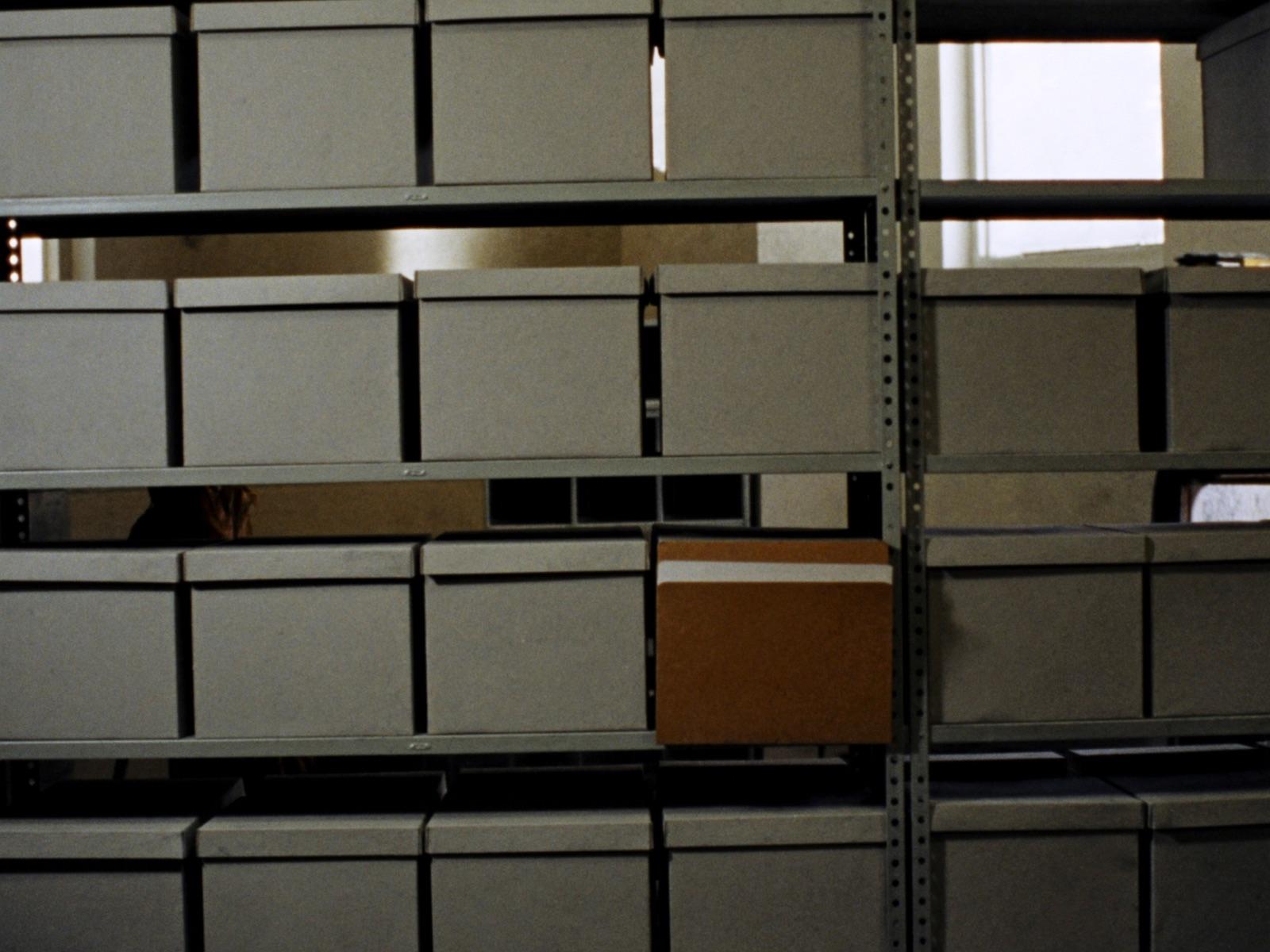 Une salle d'archives : des rangées de boîtes grises sur des rayonnages en acier, un dossier à demi sorti.