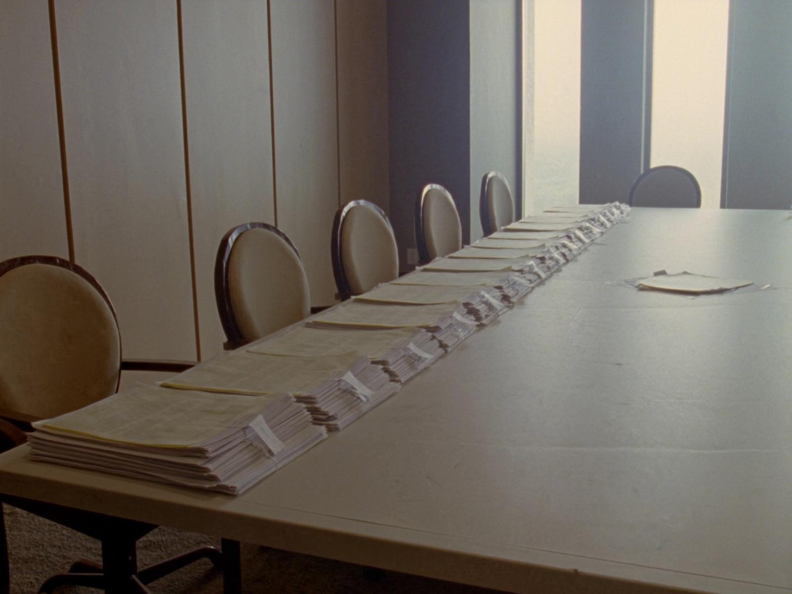 Une table de réunion avec une suite de piles de documents alignées vers les fenêtres.